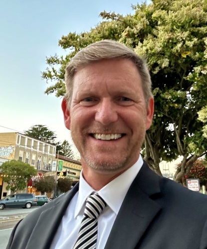 Man in a suit smiling outdoors during the daytime.